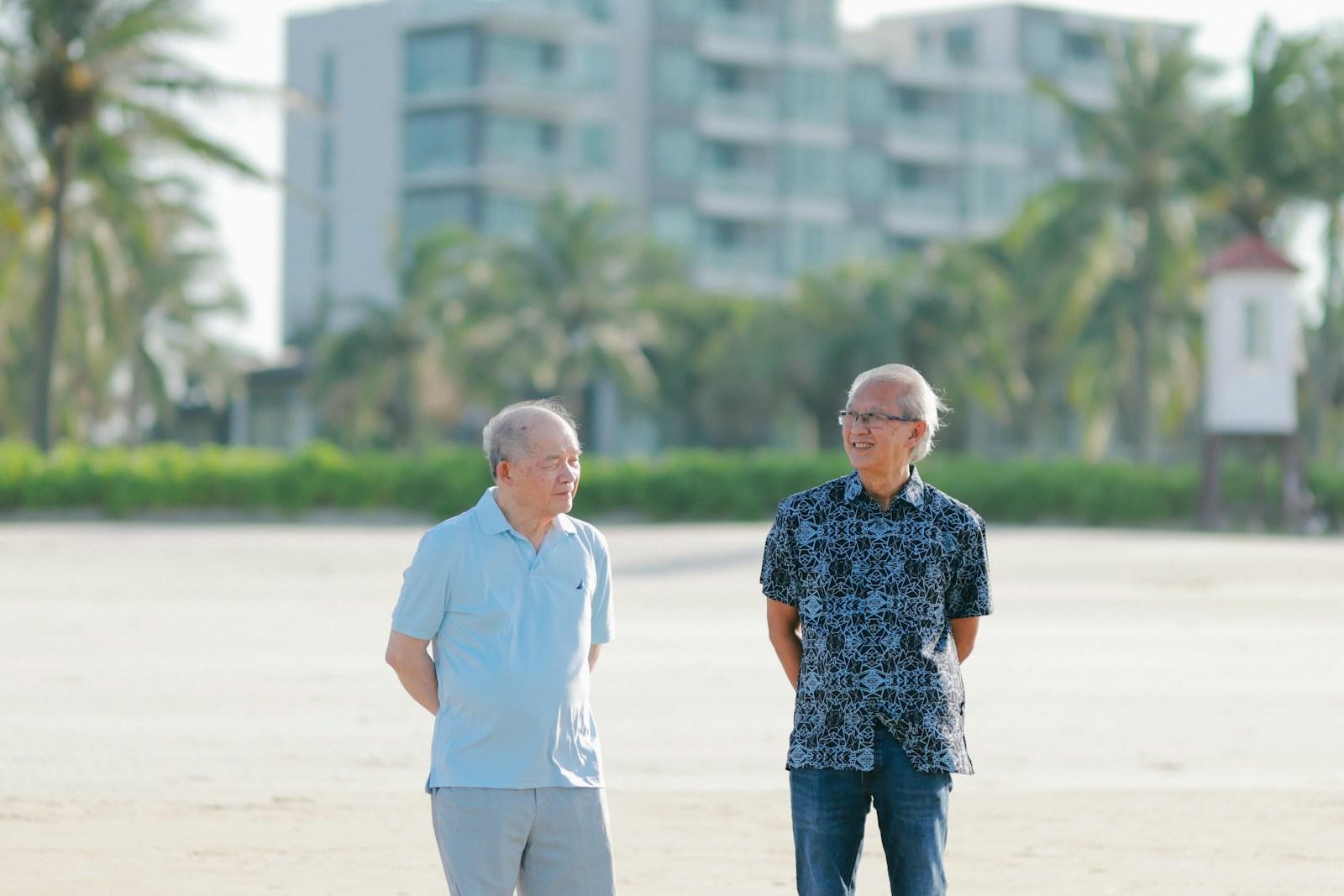 Two elderly men stand on a sandy beach.